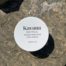 Load image into Gallery viewer, Kavana's Phyto-5 Afterglow Body Cream is light but luscious and perfectly layers under Kavana's lighweight Afterglow Body oil. Seal moisture in skin all season with these two. Seen here, the cream lies flat on a rock, opened, the cover lies flat on the upper right hand side of it. The cream is a pale ivory yellow tone. Perfect or those who intentionally seek to embody intention, using non-toxic, hormone safe, plant powered skincare.