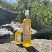 Load image into Gallery viewer, Kavana's bestselling Phyto-5 Afterglow Body oil in 30ml travel size mini glass bottle stands next to the large Afterglow Body oil- 100ml size. Both are refillable, reusable and recyclable after cleaning. The scent is very green and spa-like and uplifting. The small bottle is perfect to bring to a massage and refill with the larger one for taking on vacation as well. Seen here standing side by side on a rock, with trees and tansy in the background of a sunny sky.