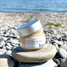 Load image into Gallery viewer, This mattifying and redness calming cream by Kavana Skincare, is pictured here with it's cap off, on a rock pile on a rocky beach, with water in the background. The Cream is slightly beigey, light ochre tinted, due to a balancing blend of Sea Buckthorn seed oil and Zinc Oxide and clays, as well as Kavana's Phyto-5 blend of plant oils.