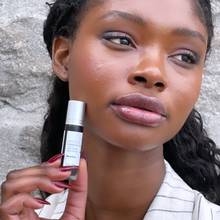 Load image into Gallery viewer, Model Raissa is seen here holding up Kavana&#39;s Phyto-5 Tinted Lip oil in LIbertine. She is wearing a striped beige jacket- with black pin stripes and is radiant in natural light. A stone wall behind her, this campaign was shot in Old MOntreal.
