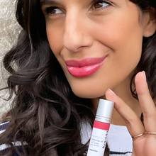 Load image into Gallery viewer, Model Kristel is seen here in tight close up, holding up a bottle of Capucine, Kavana&#39;s Phyto-5 TInted lip oil, which is a rich coral shade. She wears it on her full lips and has a blue and white mariniere sweater on, with brown hair cascading curls over it.
