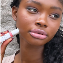 Load image into Gallery viewer, Model Raissa is seen here wearing a sheer tint in Coraline by Kavana. She is holding the frosted glass roller bottle of lip oil up to her cheek and looking to the side away from it.
