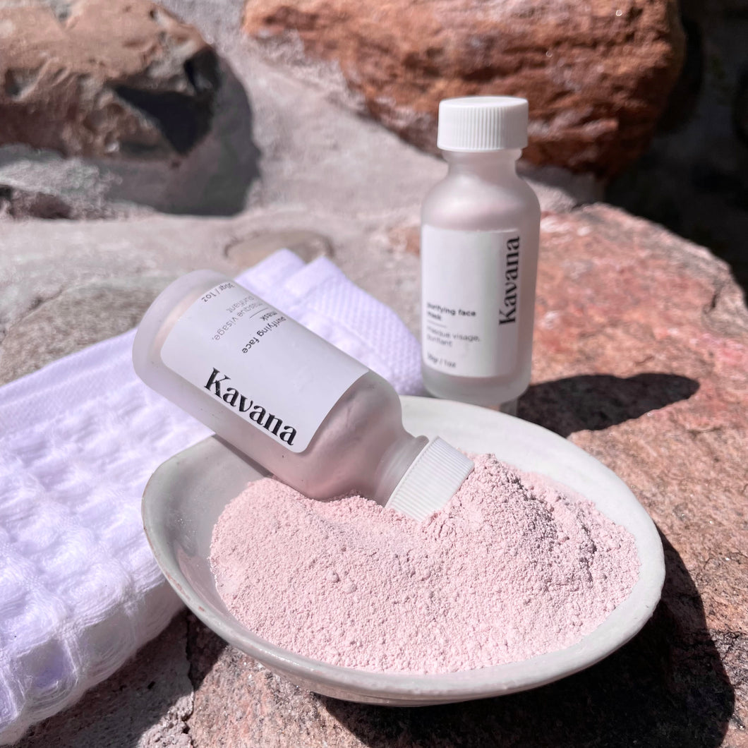 Kavana's Purifying powder mask, is seen here: a 1oz./30ml. frosted clear glass bottle with a white cap, tilted into a ceramic ivory coloured bowl filled with the mask powder. Another bottle is in the background and a white, folded waffle towel is to the left, under the bottle that is tilting over into the bowl of powder.