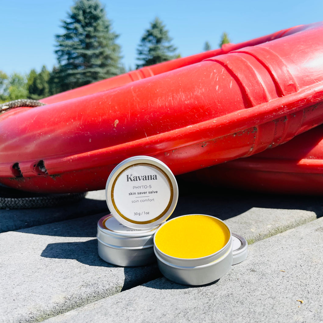 Kavana's Phyto-5 skin saver salve is perfect for late summer.  This bright yellow salve is excellent post bug bites or other broken skin. Pictured, the bright yellow salve in an open tin, with two propped salves, against a rescue buoy on a deck, with trees in the background.