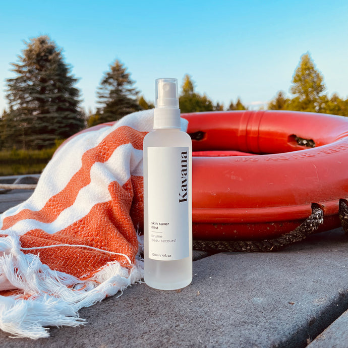 Kavana Skin Saver Mist/ Spray is great for bug bites, scars, bruises and scrapes. This clear, frosted glass bottle with a white plastic mister, is refillable and reusable. It is standing straight on a dock by a pond with trees in the bckground, propped up in front of a buoy and a Turkish towel. 