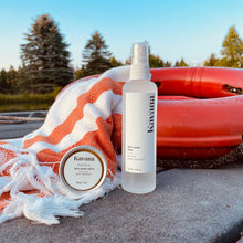 Load image into Gallery viewer, Kavana Skin Saver Mist/ Spray is great for bug bites, scars, bruises and scrapes. This clear, frosted glass bottle with a white plastic mister, is refillable and reusable. It is standing straight on a dock by a pond with trees in the bckground, propped up in front of a buoy and a Turkish towel. 
