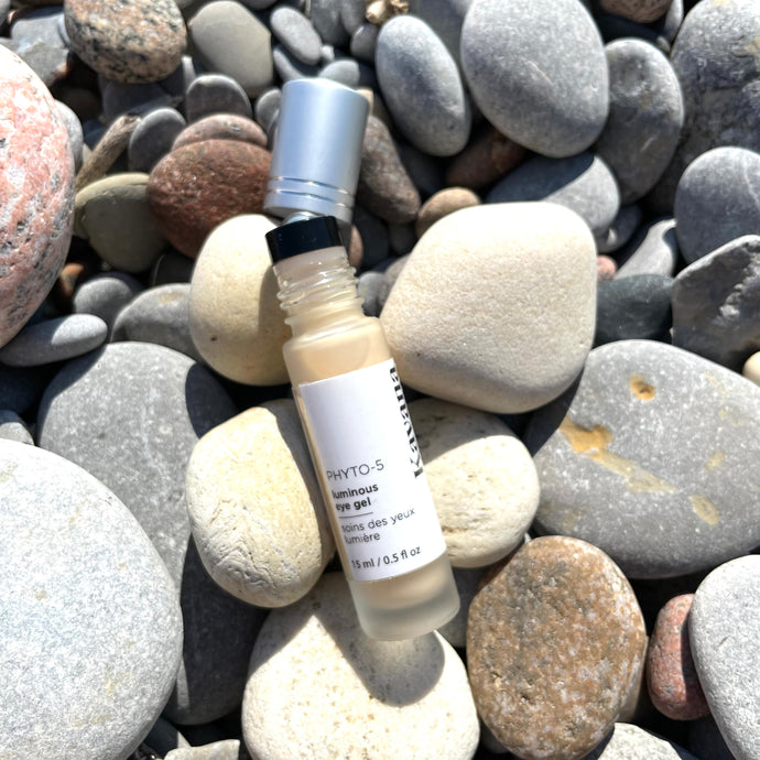 Kavana's Phyto-5 Luminous Eye Gel, is seen here opened, with roller ball top showing and cap just off the bottle to the right , on a bed of stones at the beach. Packed with Green Coffee extract to depuff, Hyaluronic Acid to hdyrate, Blue Cornflower to soothe and a mica blend to hilight, this is the cooling eye gel you need! It also has Kavana's signature Phyto-5 plant blend of Calendula, COmfrey, Echinacea, Plantain and Rosehip seed oil. Delighftul as a 2-in-1 multipurpose skincare and makeup product!