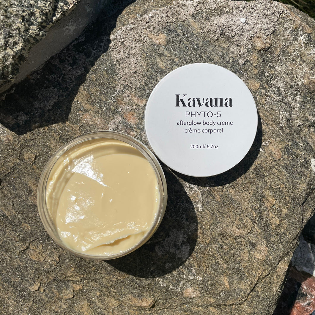 Kavana's Phyto-5 Afterglow Body Cream is light but luscious and perfectly layers under Kavana's lighweight Afterglow Body oil. Seal moisture in skin all season with these two. Seen here, the cream lies flat on a rock, opened, the cover lies flat on the upper right hand side of it. The cream is a pale ivory yellow tone. Perfect or those who intentionally seek to embody intention, using non-toxic, hormone safe, plant powered skincare.