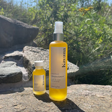 Load image into Gallery viewer, Kavana&#39;s bestselling Phyto-5 Afterglow Body oil in 30ml travel size mini glass bottle stands next to the large size 100ml bottle. Both are refillable, reusable and recyclable after cleaning. The scent is very green and spa like and uplifting. The small bottle is perfect to bring to a massage and refill with the larger one for taking on vacation as well. Seen here standing side by side on a rock, with trees and tansy in the background of a sunny sky. 
