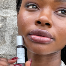 Load image into Gallery viewer, Model Raissa is seen in tight close up holding up Kavana&#39;s Phyto-5 Tinted LIp oil in Libertine- a dark black honey kind of shade. This sheer blackberry has golden undertones to it-giving it a warmer look on skin than a cool blackberry shade might be expected to.
