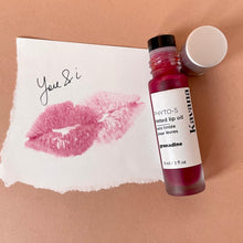 Load image into Gallery viewer, Kavana&#39;s Phyto-5 tinted lip treatment oil in shade Grenadine, is a sheer pomegranate red, berry tint for lips. Seen open over top of a love note, the roll on bottle looks like it was just used. The note says u &amp; i on it with a lip print in the colour. 

